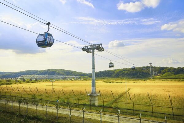 A photo of the Paju DMZ Gondola crossing the Imjin River during Autumn with a bright yellow coloured field underneath and barbed wire surrounding the river